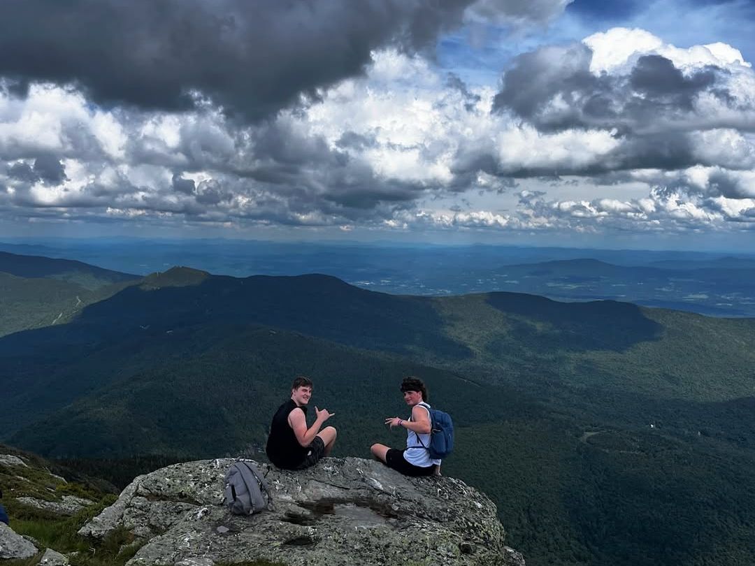 A breathtaking view from the summit of Mount Mansfield, Vermont’s highest peak, shows two hikers resting atop rugged rocks with sweeping views of the lush Green Mountains below. Dark, dramatic clouds cast shadows over the valleys, creating a striking scene of adventure and serenity.