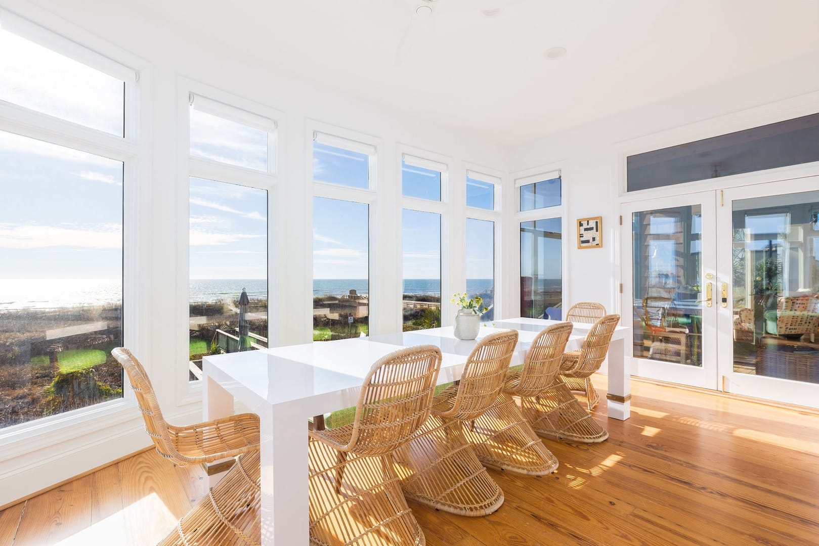 A bright dining room filled with windows looks out over the ocean and dunes. The white table and woven chairs give the space a warm, coastal feel. Sunlight fills every corner, making it a cheerful spot for meals and ocean views.