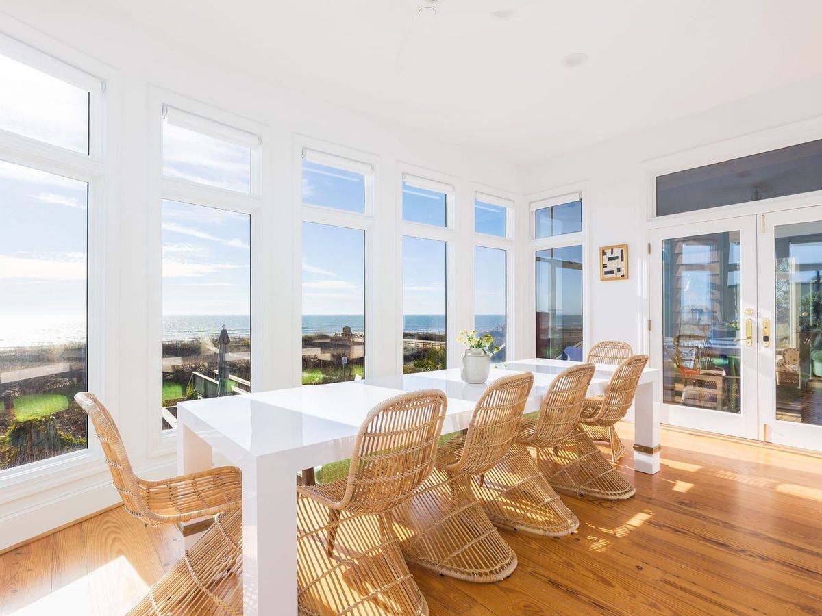 A bright dining room filled with windows looks out over the ocean and dunes. The white table and woven chairs give the space a warm, coastal feel. Sunlight fills every corner, making it a cheerful spot for meals and ocean views.