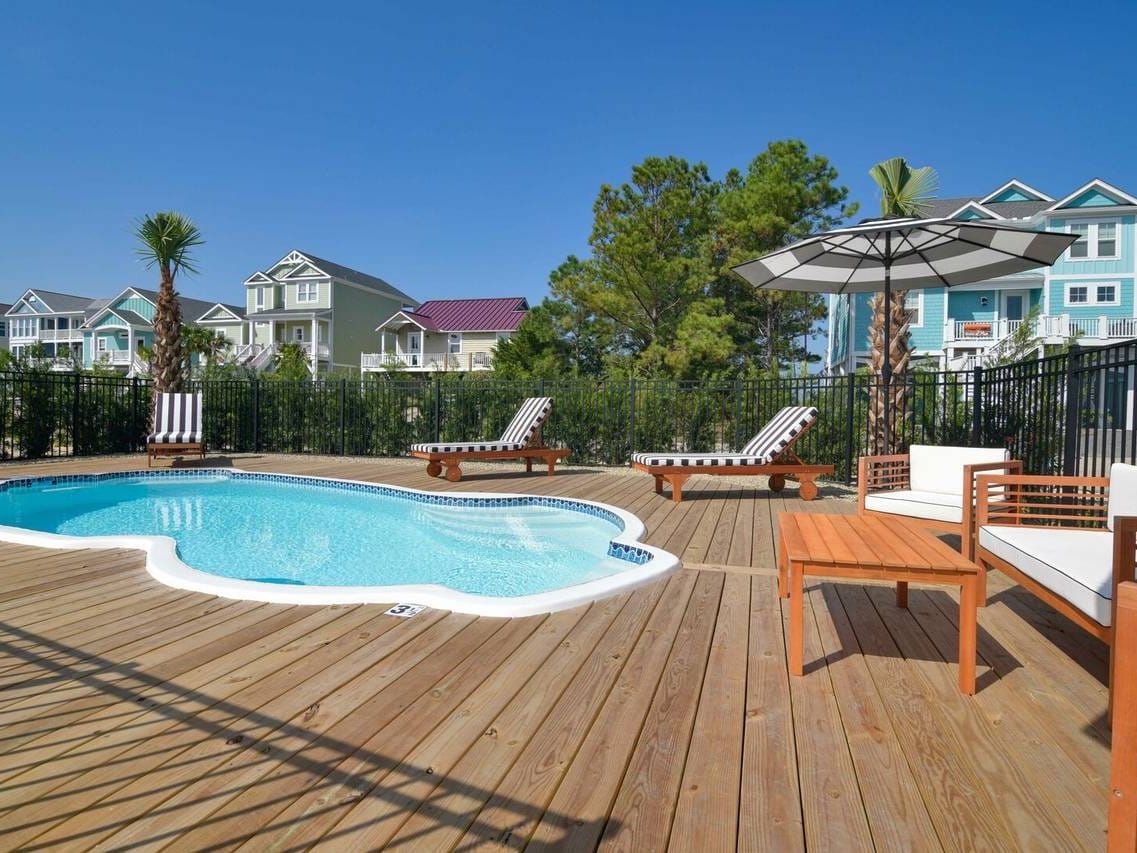 A private pool surrounded by a wood deck, striped loungers, and palm trees in a beachside neighborhood. Perfect for relaxing days under the sun in Holden Beach.