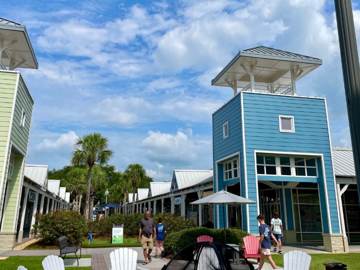 A colorful outdoor shopping village with pastel buildings, palm trees, Adirondack chairs, and people walking along a sunny pathway under a bright blue sky with scattered clouds.