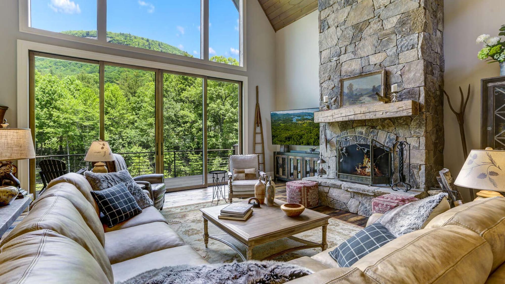 This mountain home features a spacious living room with a tall stone fireplace and floor-to-ceiling windows overlooking lush green hills. The open layout and natural light create a comfortable setting for a North Carolina mountain stay. It’s perfect for relaxing after a day of hiking or exploring.
