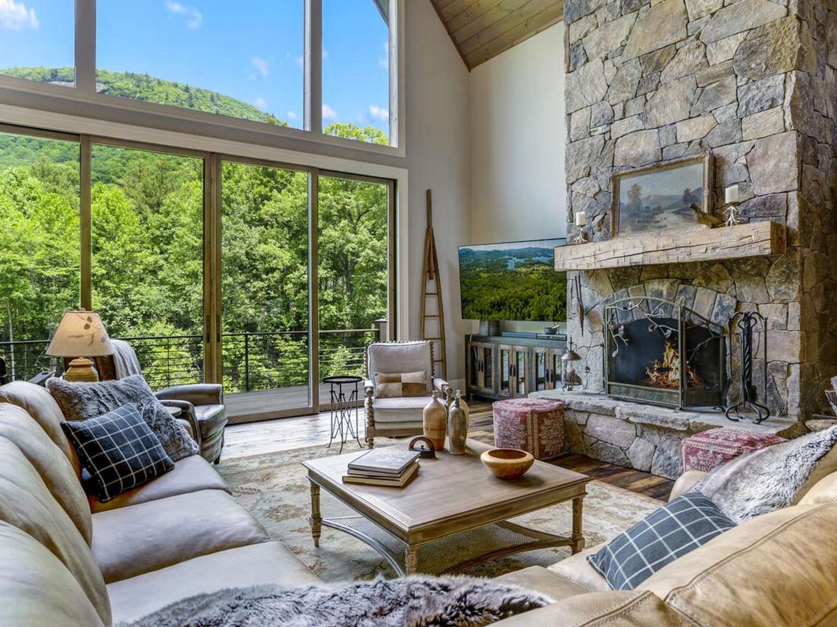 This mountain home features a spacious living room with a tall stone fireplace and floor-to-ceiling windows overlooking lush green hills. The open layout and natural light create a comfortable setting for a North Carolina mountain stay. It’s perfect for relaxing after a day of hiking or exploring.