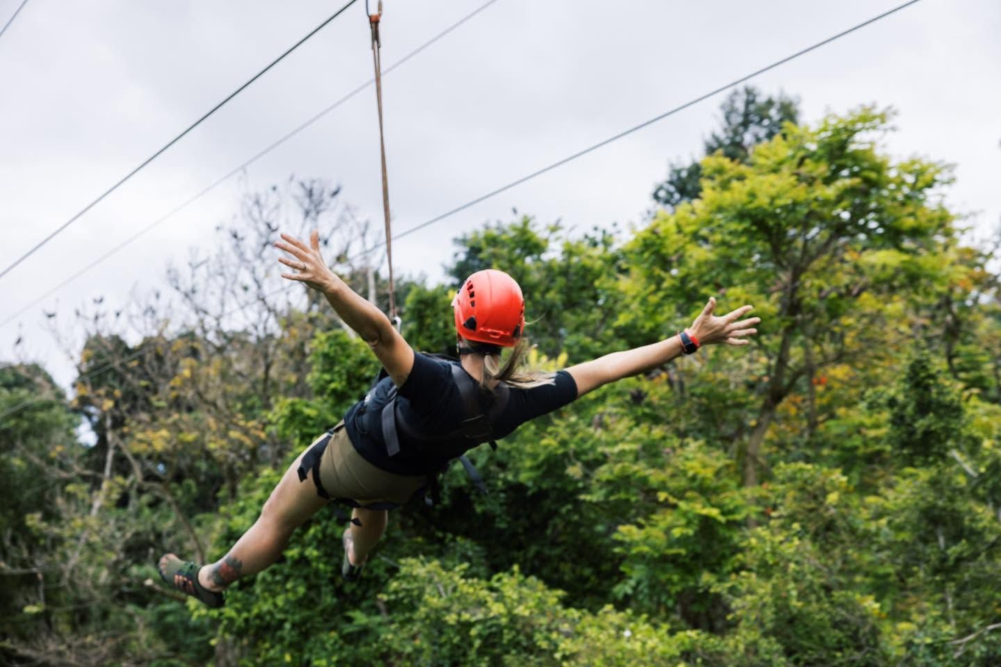 A woman in a red helmet spreads her arms wide while ziplining through Kauai's lush tropical forest canopy on an Outfitters Kauai zipline adventure tour. 