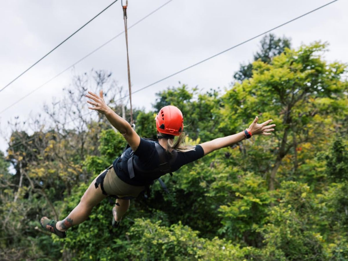 A woman in a red helmet spreads her arms wide while ziplining through Kauai's lush tropical forest canopy on an Outfitters Kauai zipline adventure tour.