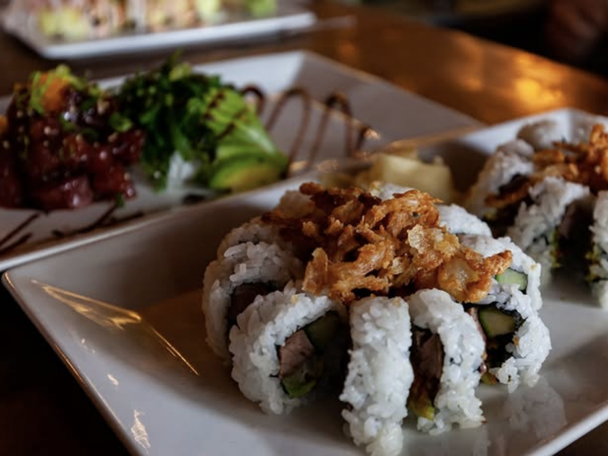 Close-up of sushi rolls topped with crispy fried onions, served alongside fresh poke with avocado slices and a drizzle of dark sauce on a rectangular white plate.