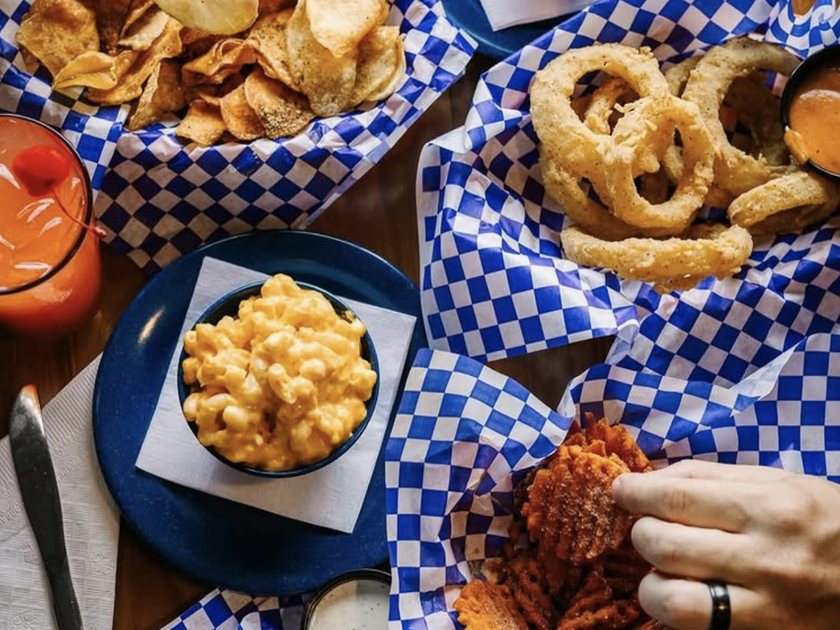 A table filled with comfort food appetizers including baskets of homemade potato chips, onion rings with dipping sauce, waffle fries, and a bowl of creamy mac and cheese, alongside a bright red cocktail with a cherry garnish.