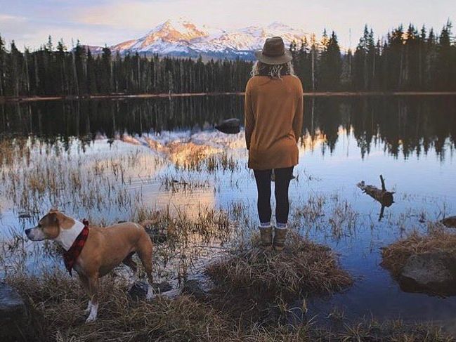 A woman and her dog stand at the edge of a calm alpine lake, gazing at snow-capped mountains reflecting in the water. The tranquil moment captures the rugged beauty of Oregon’s Three Sisters Wilderness.