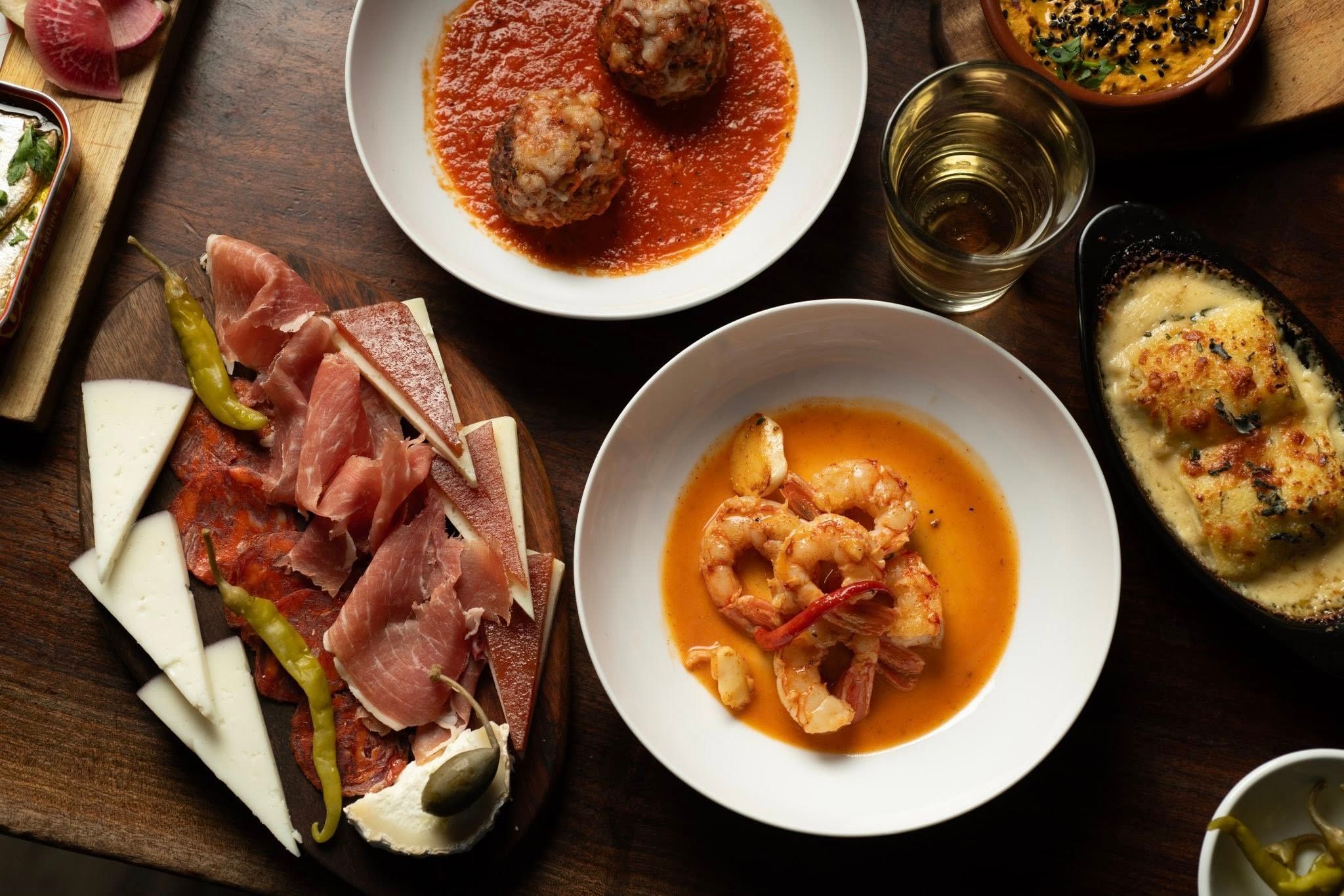An overhead spread of Spanish tapas covers a rustic wooden table, featuring a charcuterie board with jamón, chorizo, manchego, and pickled peppers, alongside garlic shrimp in golden sauce, meatballs in tomato sauce, and a bubbling baked dish. A glass of sherry and small bowls of dips complete the table. 