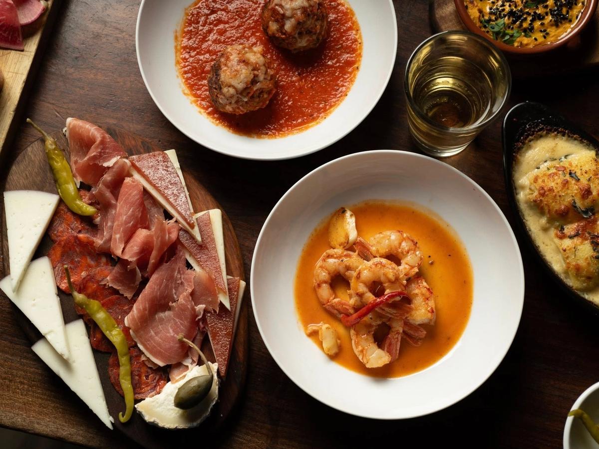 An overhead spread of Spanish tapas covers a rustic wooden table, featuring a charcuterie board with jamón, chorizo, manchego, and pickled peppers, alongside garlic shrimp in golden sauce, meatballs in tomato sauce, and a bubbling baked dish. A glass of sherry and small bowls of dips complete the table.