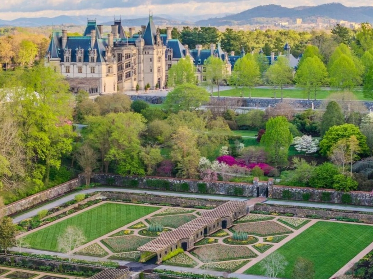 Aerial view of a grand historic estate surrounded by manicured gardens, lush trees, and mountain scenery under a partly cloudy sky