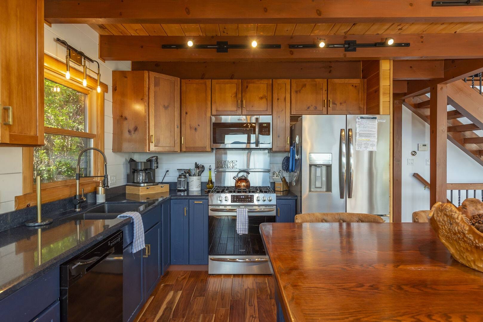 A warm, rustic-modern kitchen featuring exposed wood beams, honey-toned upper cabinets, navy blue lower cabinets, stainless steel appliances, black countertops, and a farmhouse sink beside a sunlit window, anchored by a solid wood dining table in the foreground.