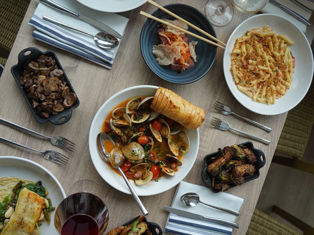 A delicious overhead view of shared plates including clams in red sauce, mac and cheese, crispy mushrooms, and toasted bread. Ideal for a laid-back meal with friends.