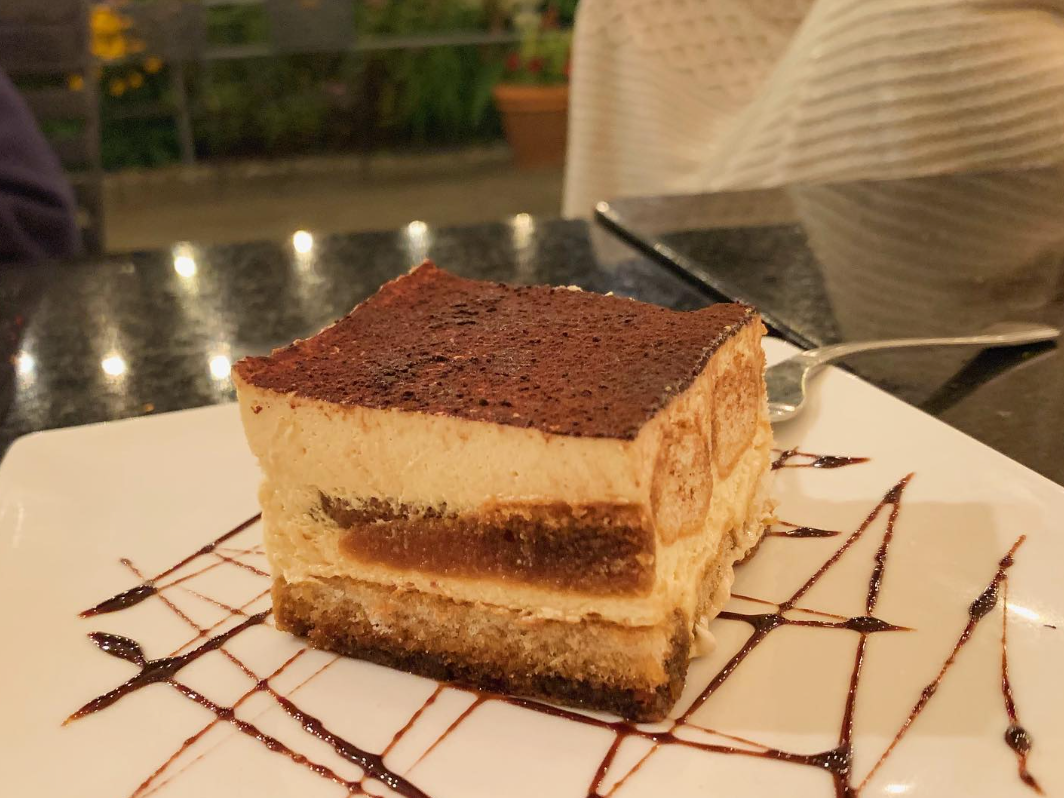 A decadent slice of tiramisu rests on a white plate with a chocolate drizzle design, set on an outdoor table under cozy evening lights