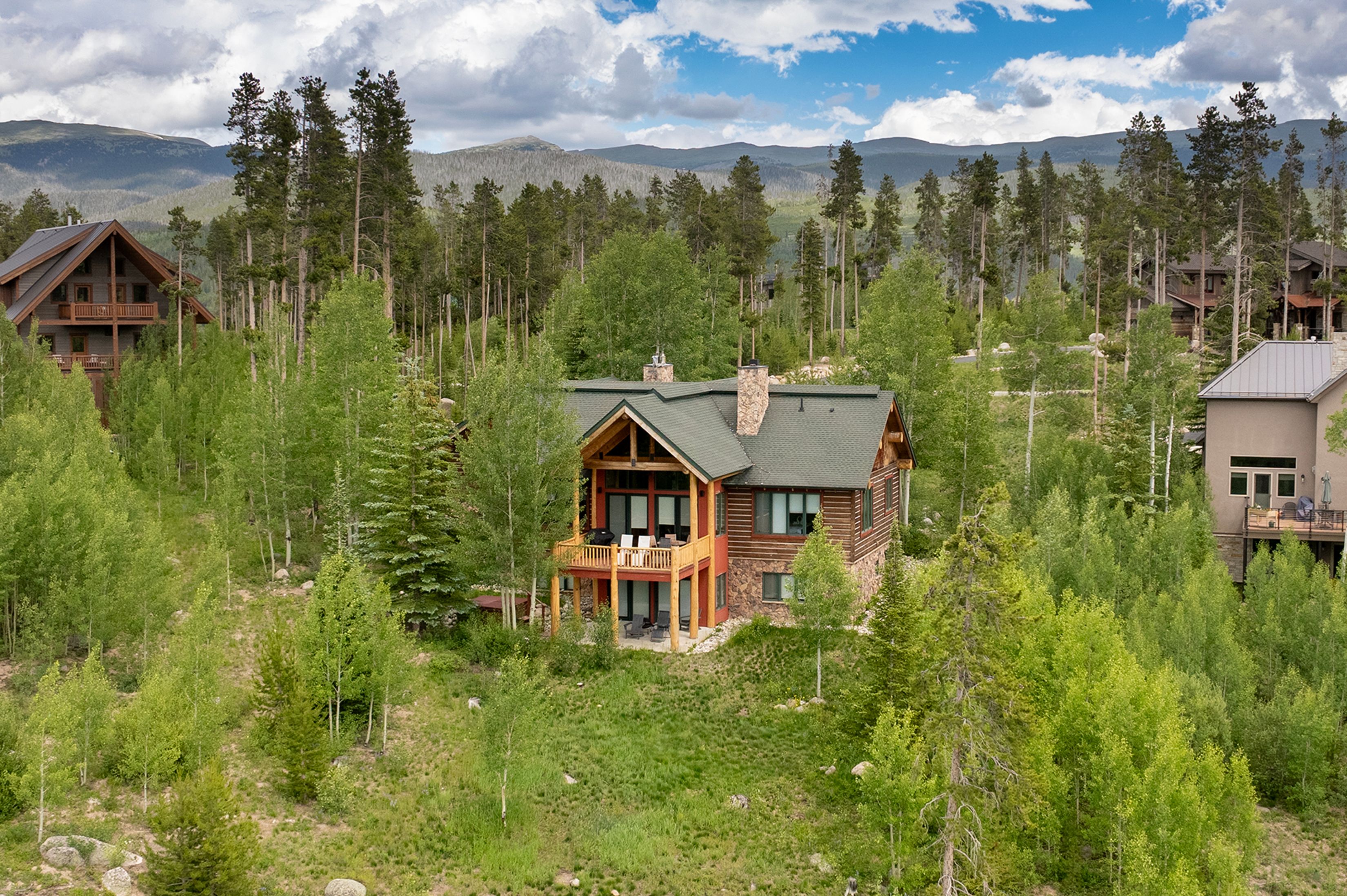 A rustic mountain cabin with a green roof and wood-and-stone exterior sits nestled in a lush forest clearing, surrounded by tall trees and other cabins with mountains in the distance under a partly cloudy sky.