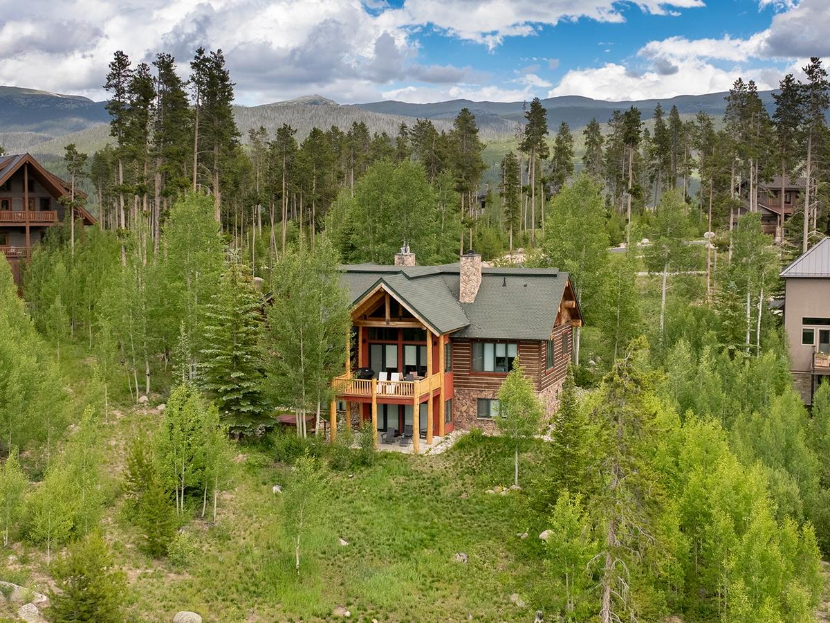 A rustic mountain cabin with a green roof and wood-and-stone exterior sits nestled in a lush forest clearing, surrounded by tall trees and other cabins with mountains in the distance under a partly cloudy sky.