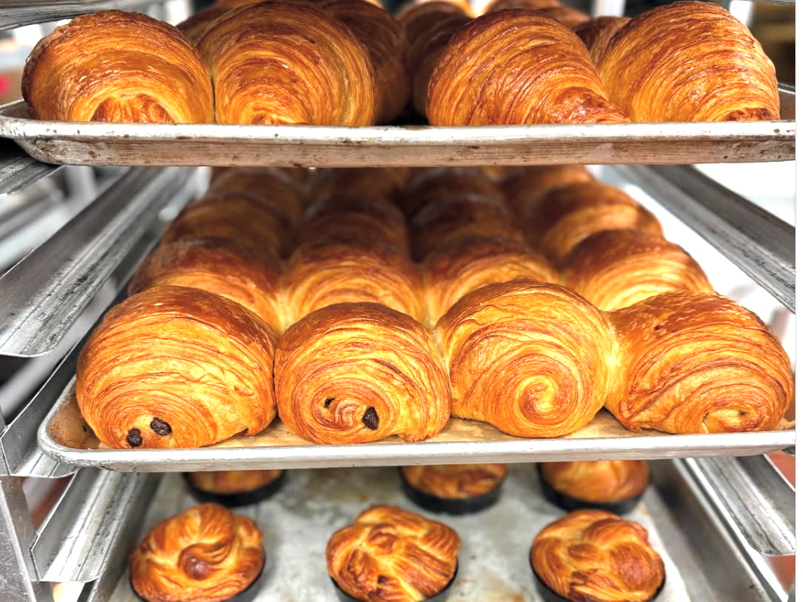 Trays of freshly baked pastries, including golden-brown croissants, pain au chocolat, and round laminated pastries cooling on bakery racks.