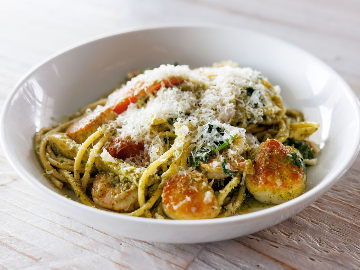 Scallop pesto linguine topped with grated parmesan and fresh tomato slices, served in a wide white bowl. This creamy seafood pasta from Blue Moon Beach Grill is rich, savory, and packed with flavor.