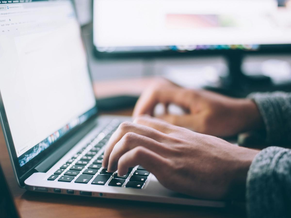 A person types on a laptop keyboard in a workspace with a large monitor in the background, suggesting focused work or digital communication.