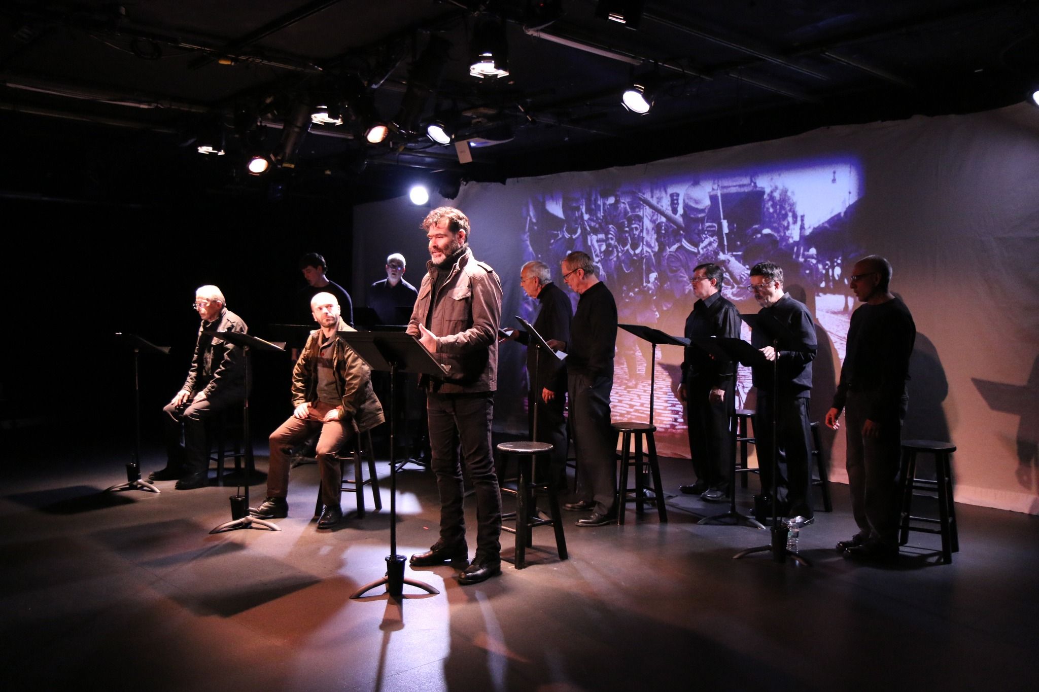 A group of actors performs a dramatic reading on a small, dimly lit stage, creating an intimate and emotional scene. The cast stands at music stands while a historic war image is projected behind them, adding depth to the performance. The warm lights and simple setup make the moment feel raw and powerful.