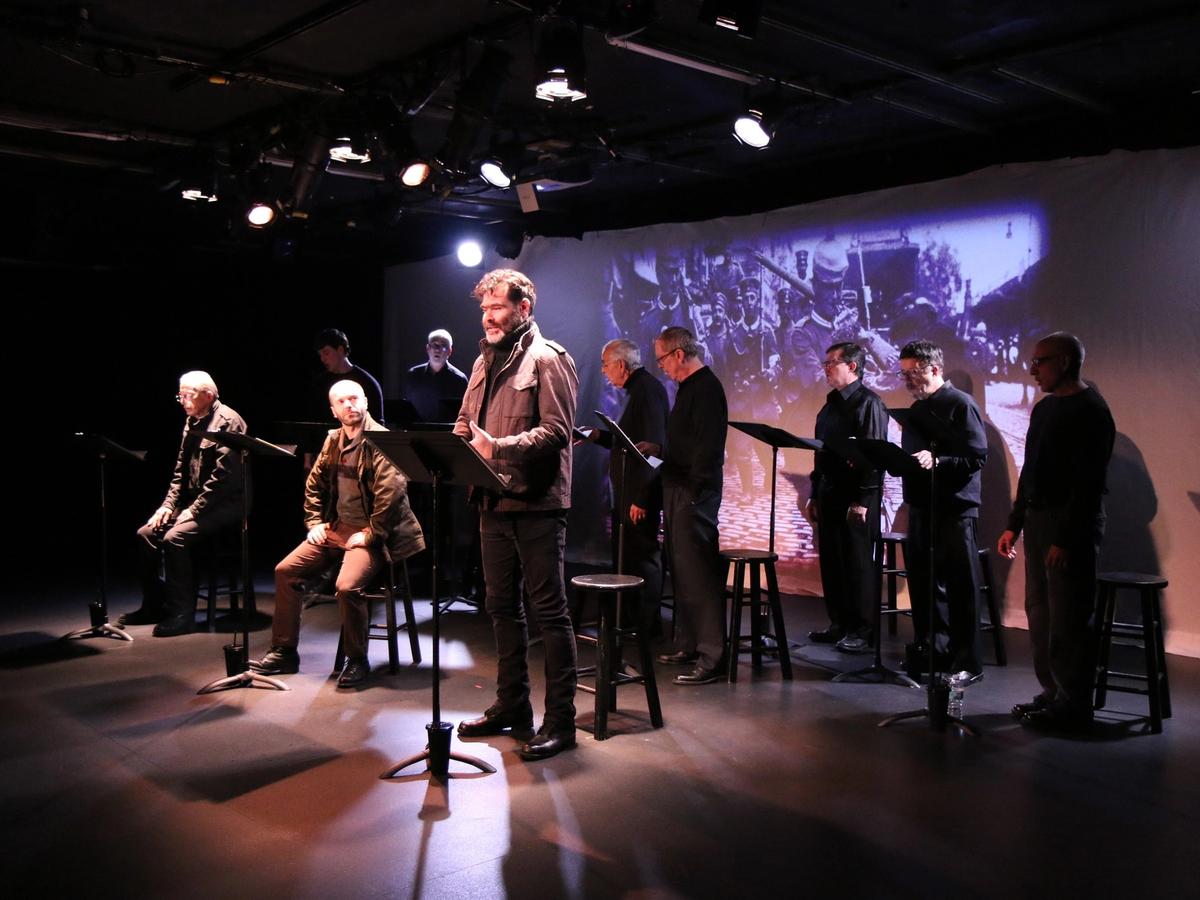 A group of actors performs a dramatic reading on a small, dimly lit stage, creating an intimate and emotional scene. The cast stands at music stands while a historic war image is projected behind them, adding depth to the performance. The warm lights and simple setup make the moment feel raw and powerful.