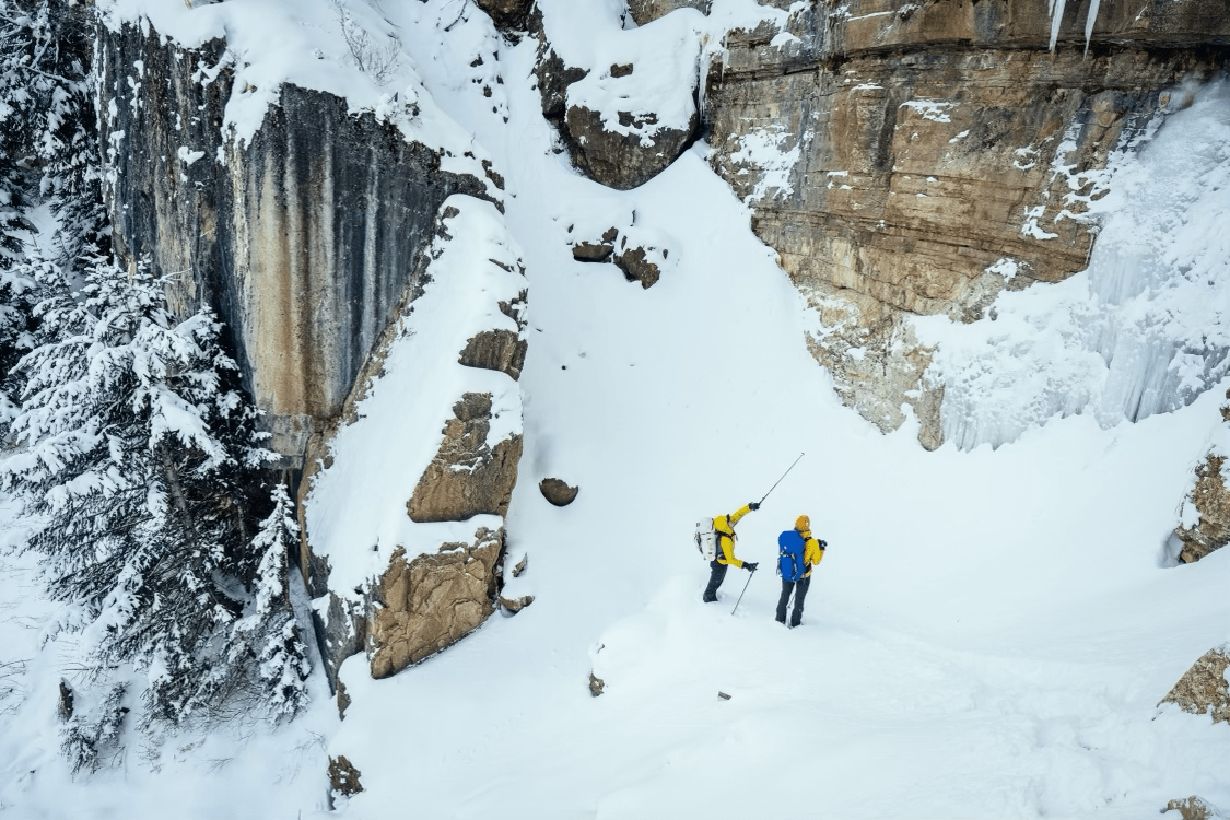 Two climbers dressed in bright gear stand below an icy cliffside, pointing out their route before beginning a challenging winter ascent. The snow-covered canyon and frozen waterfalls make for a breathtaking Wyoming adventure.