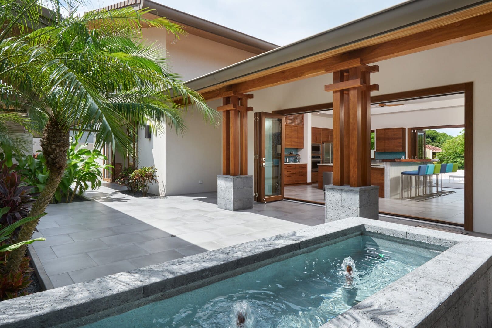Open-air courtyard with a small stone water feature, tropical plants, and wide wooden-framed doors leading into a modern kitchen and dining area.