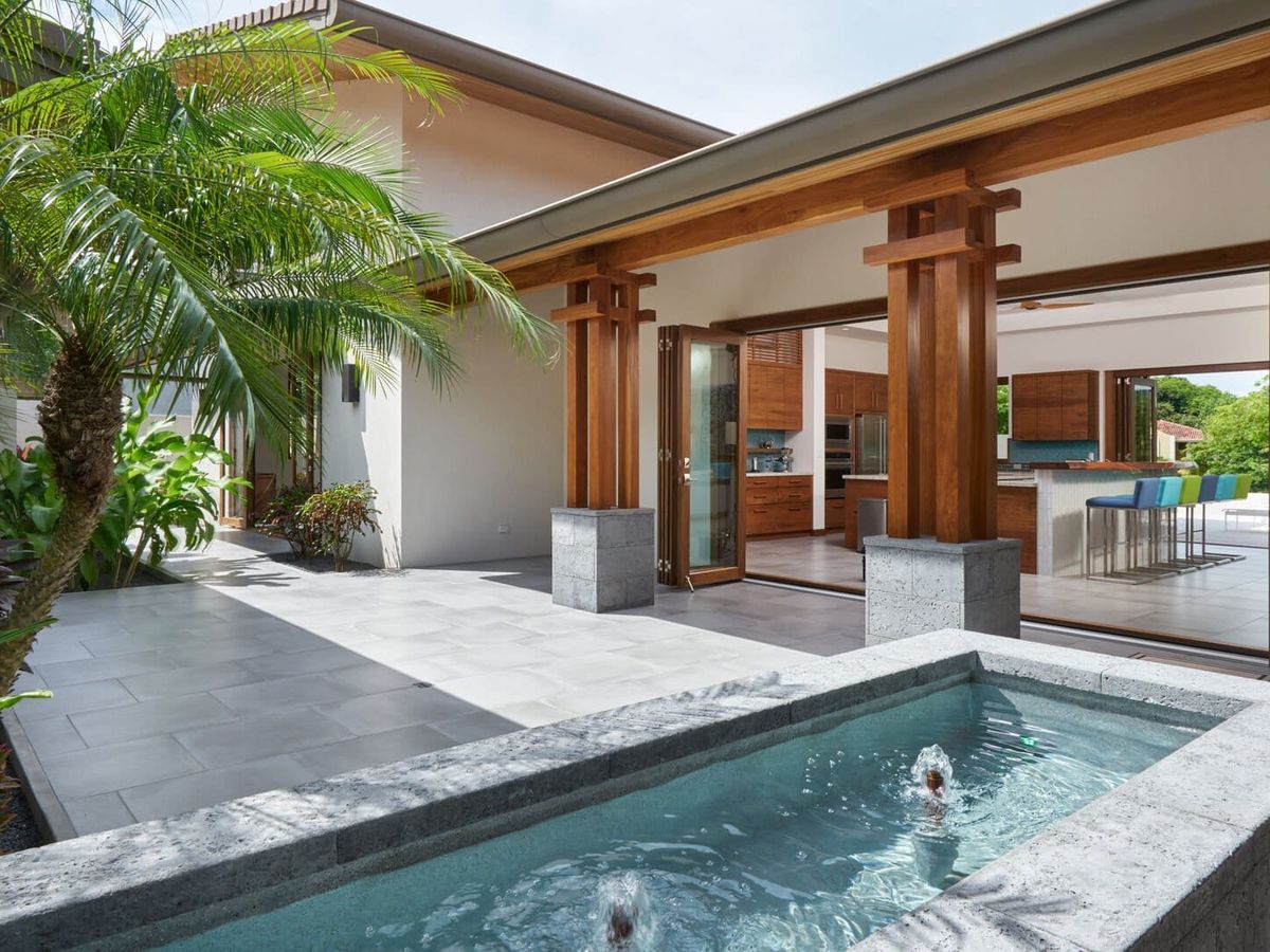 Open-air courtyard with a small stone water feature, tropical plants, and wide wooden-framed doors leading into a modern kitchen and dining area.