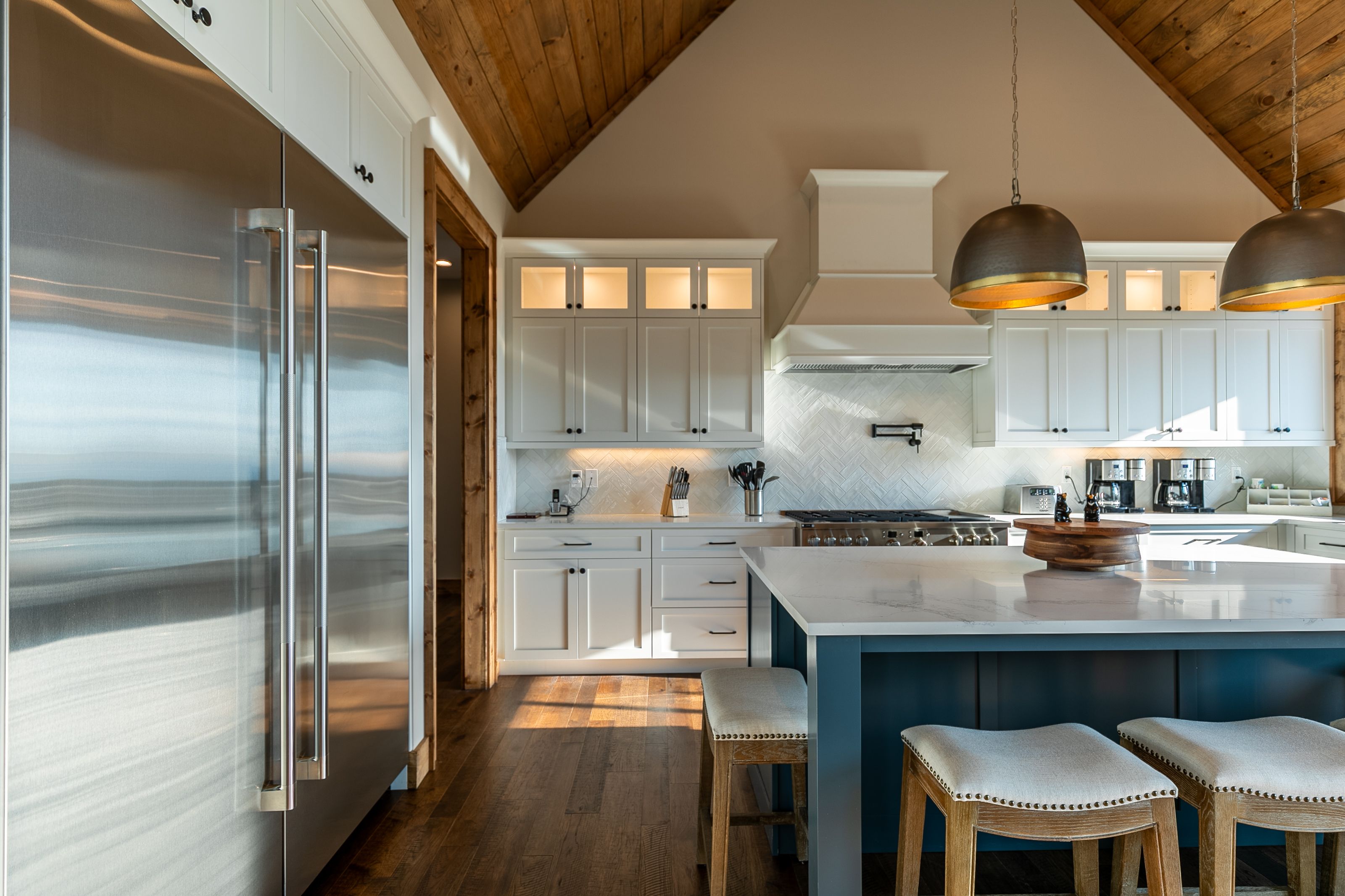 "A spacious kitchen with a vaulted wooden ceiling features white cabinetry, a blue island with a marble countertop, and a large stainless steel refrigerator. Pendant lighting hangs over the island, complementing the rustic wood flooring. The space includes a gas stove with a white range hood, a built-in pot filler, and a coffee station.