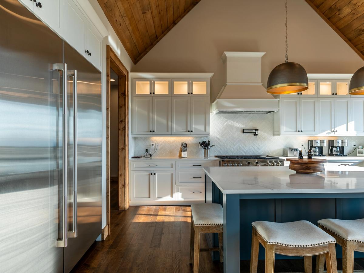 "A spacious kitchen with a vaulted wooden ceiling features white cabinetry, a blue island with a marble countertop, and a large stainless steel refrigerator. Pendant lighting hangs over the island, complementing the rustic wood flooring. The space includes a gas stove with a white range hood, a built-in pot filler, and a coffee station.