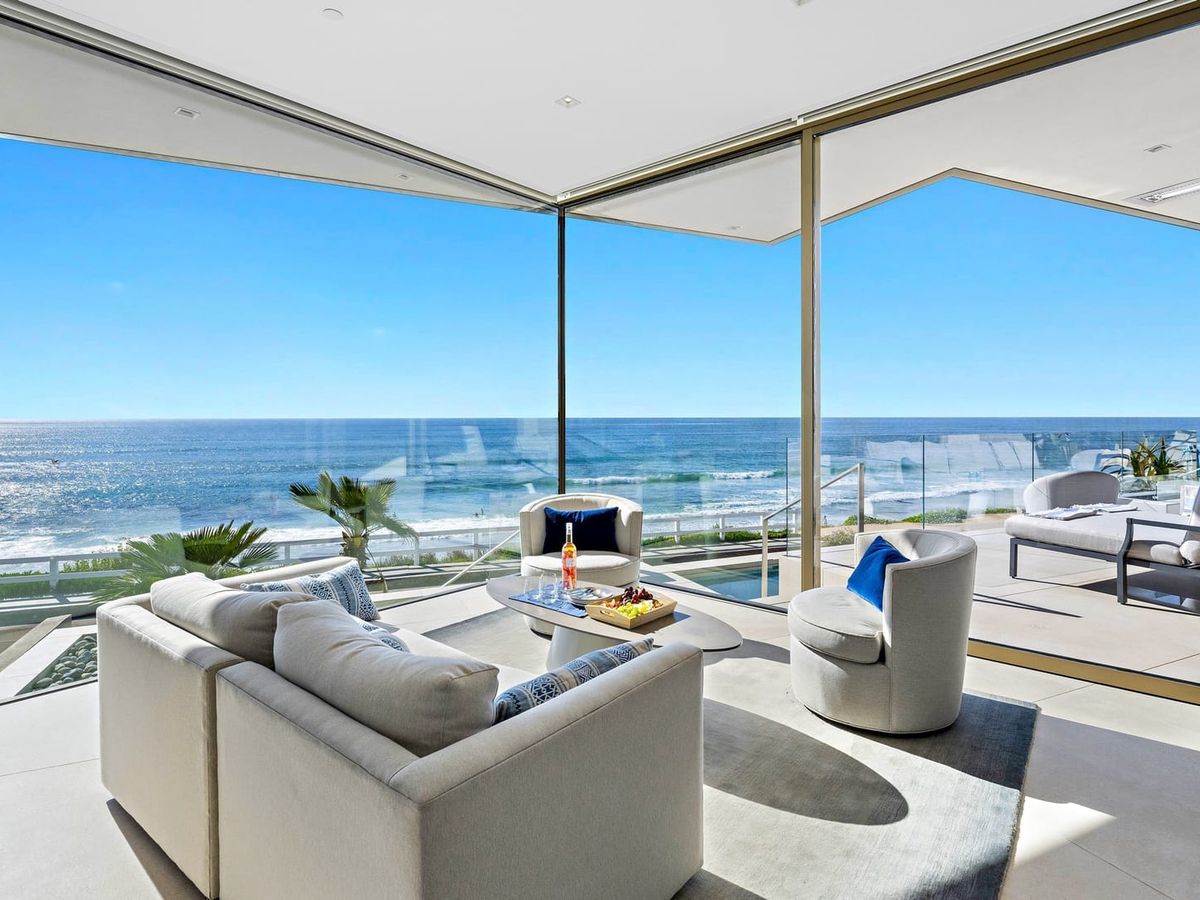 A modern living room with floor-to-ceiling windows shows a stunning view of the bright blue ocean. Sunlight reflects off the water, filling the space with light. It’s a relaxing setting for enjoying the waves all day.