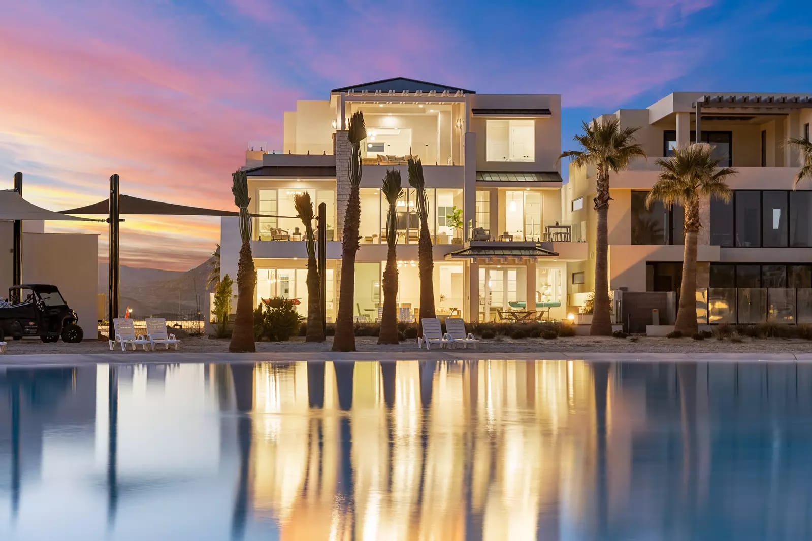 A modern beachfront villa glows warmly at sunset, with tall palm trees in front and its lights reflecting across a calm pool of water.