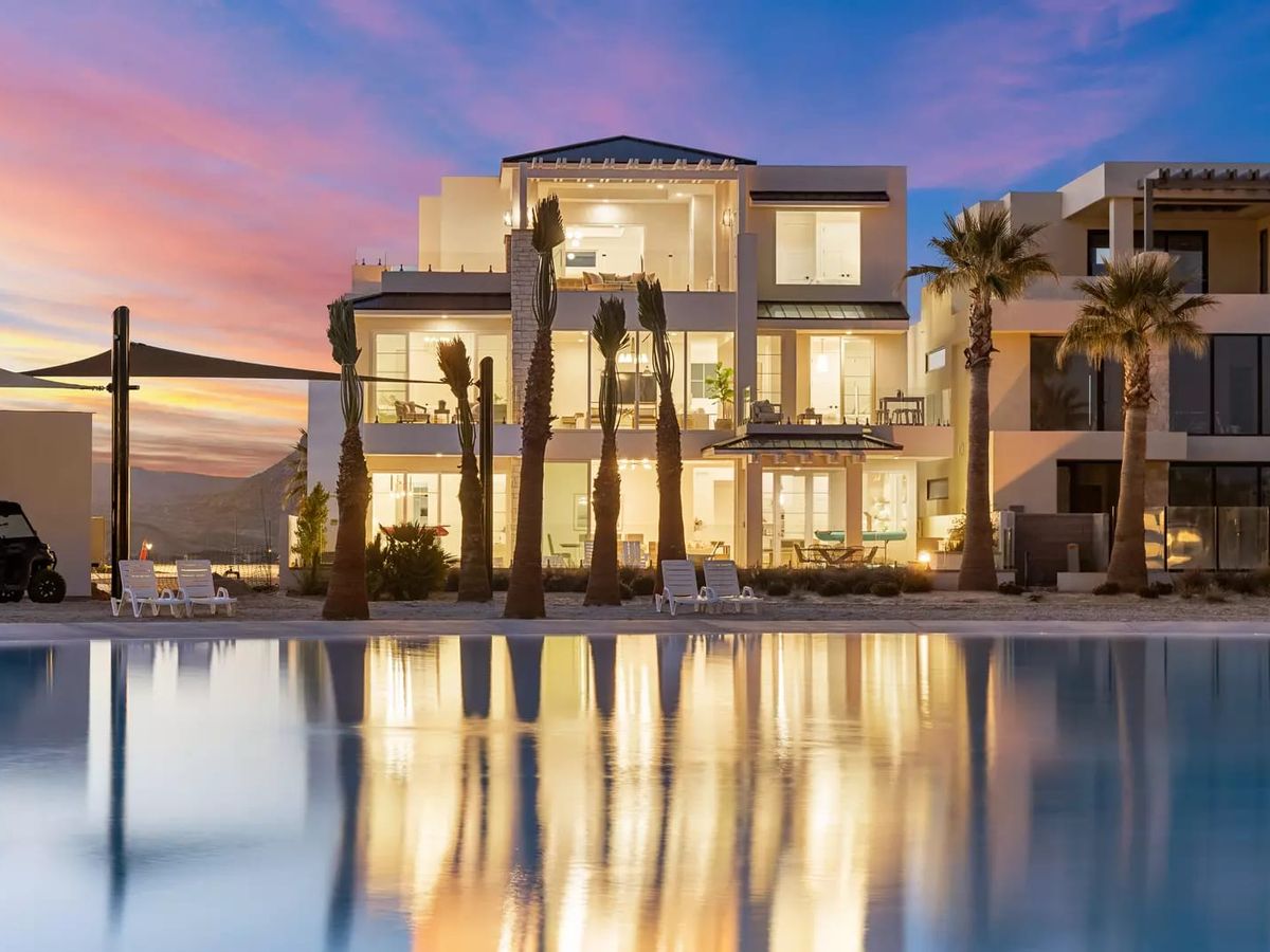 A modern beachfront villa glows warmly at sunset, with tall palm trees in front and its lights reflecting across a calm pool of water.