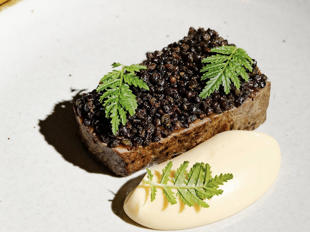 A refined dish featuring a rectangular slice of meat topped with glossy black lentils or caviar and garnished with fresh green herbs, served alongside a quenelle of creamy mashed potatoes or purée on a minimalist white plate.