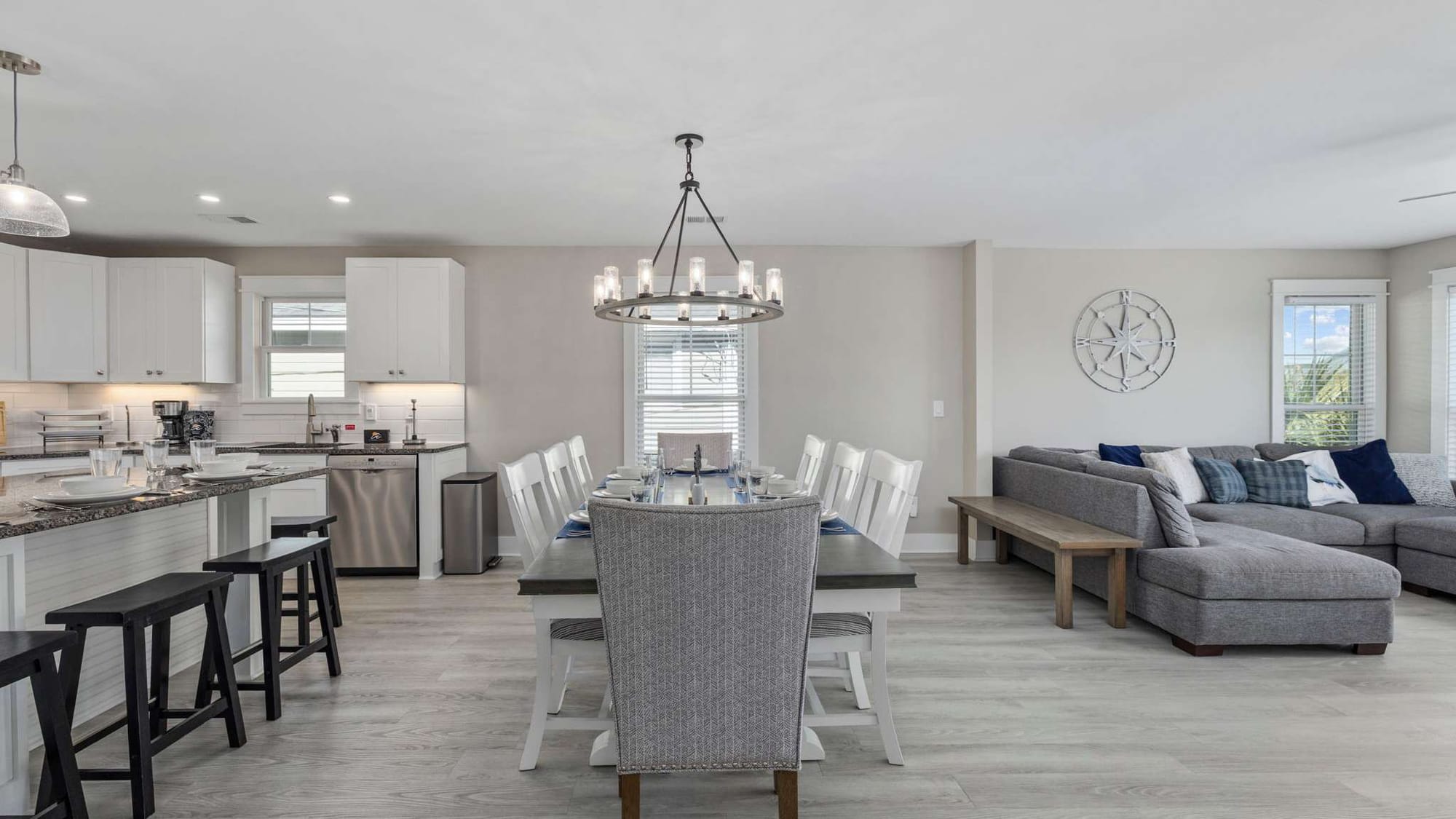 A bright open-concept living, dining, and kitchen area at Beach Days and Fairways vacation rental features a large farmhouse dining table set for ten beneath a wagon wheel chandelier, white shaker cabinets, and a coastal compass rose wall accent. Light wood floors, navy blue accents, and a spacious sectional sofa complete the welcoming beach house aesthetic.