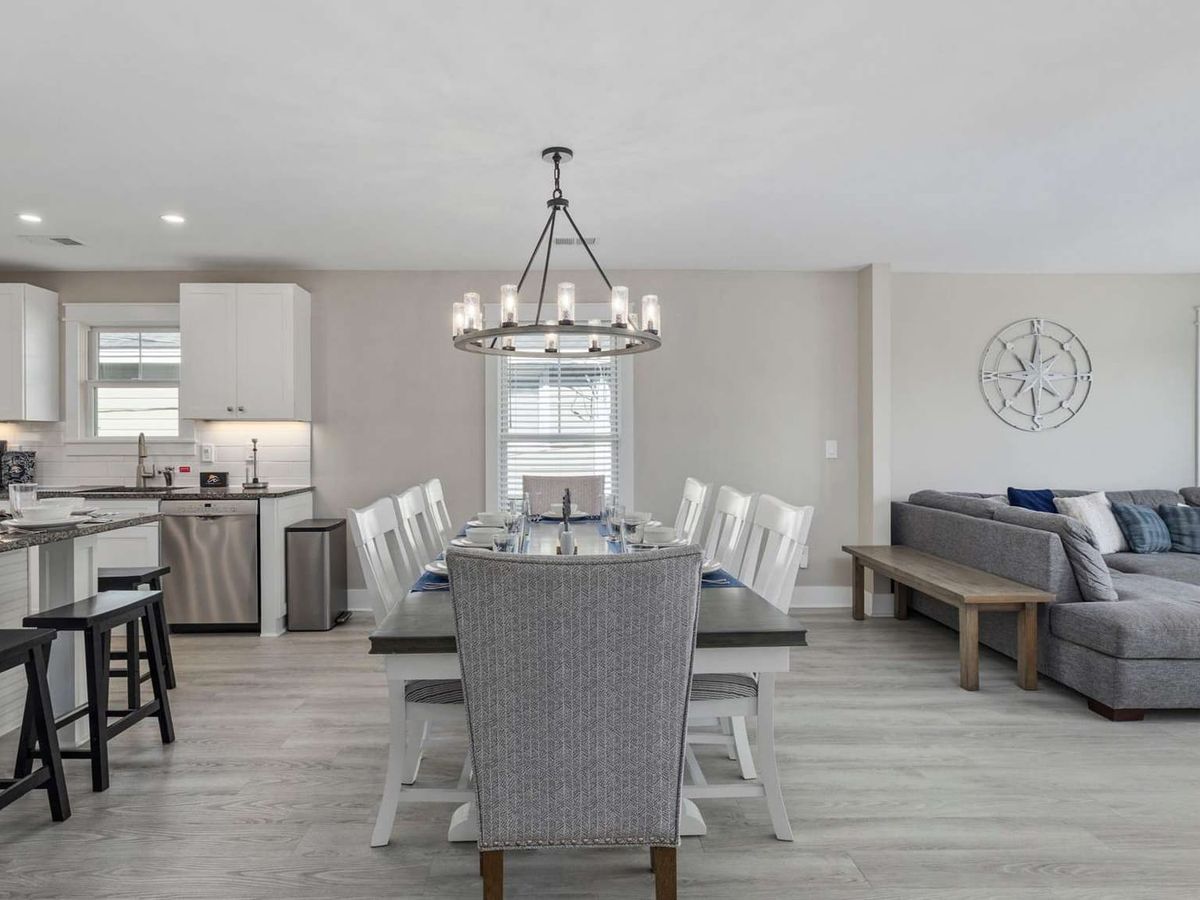 A bright open-concept living, dining, and kitchen area at Beach Days and Fairways vacation rental features a large farmhouse dining table set for ten beneath a wagon wheel chandelier, white shaker cabinets, and a coastal compass rose wall accent. Light wood floors, navy blue accents, and a spacious sectional sofa complete the welcoming beach house aesthetic.