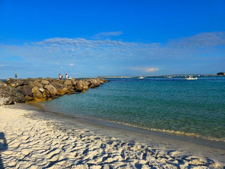 A peaceful view of O’Steen Public Beach Access with white sandy shores, clear turquoise water, and a rocky jetty where people enjoy fishing under a bright blue sky. Perfect for a quiet beach day in Destin, Florida.