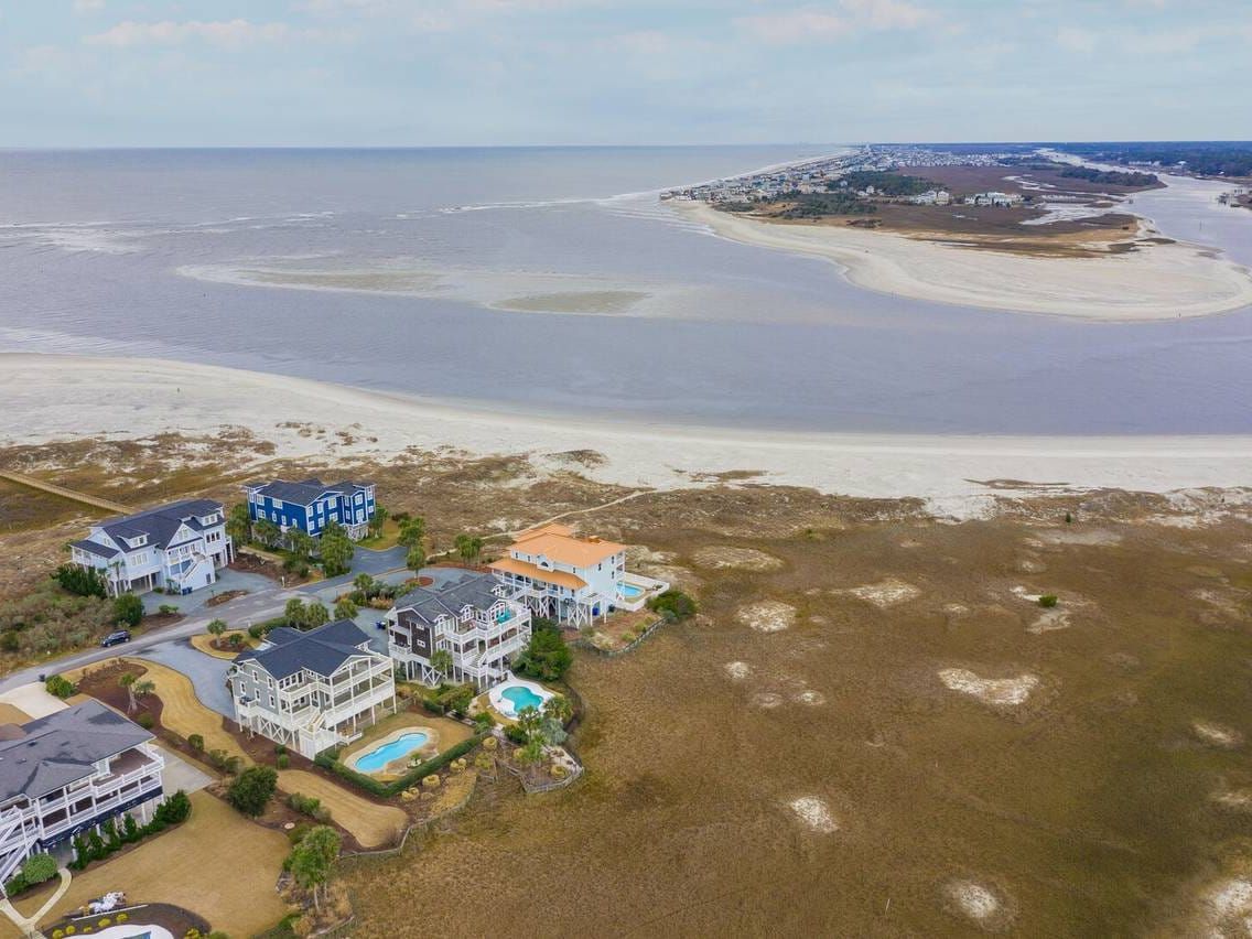 Aerial view of beachfront vacation homes with pools along a sandy coastline, overlooking a calm inlet and barrier island in the distance.