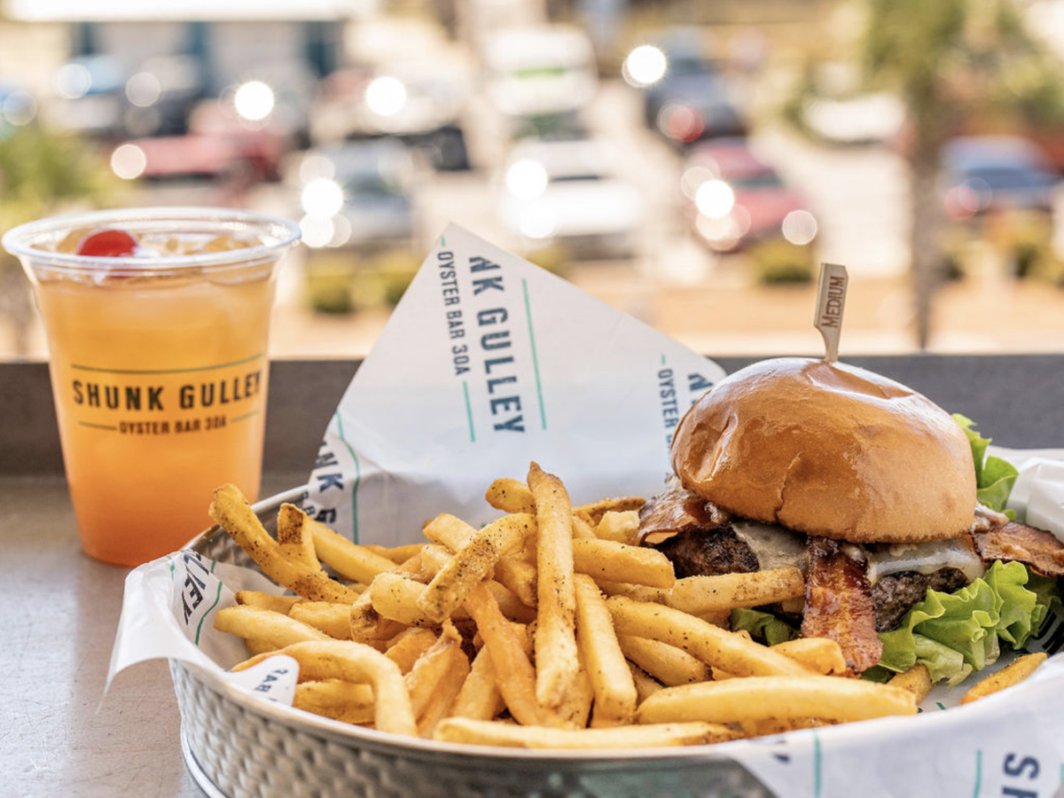 A bacon cheeseburger with lettuce served alongside seasoned fries in a metal basket, paired with a tropical cocktail from Shunk Gulley Oyster Bar 30A.