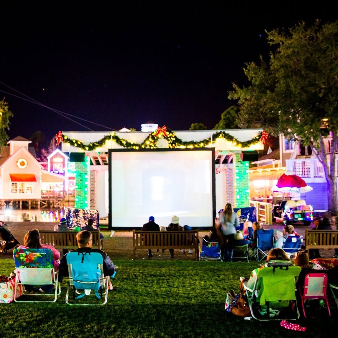 Families sit on lawn chairs and blankets while watching an outdoor holiday movie on a large screen at night. Festive lights, Christmas garlands, and nearby shops create a warm and cheerful setting for a community movie night. This image captures the cozy feel of outdoor holiday movies with family and friends.