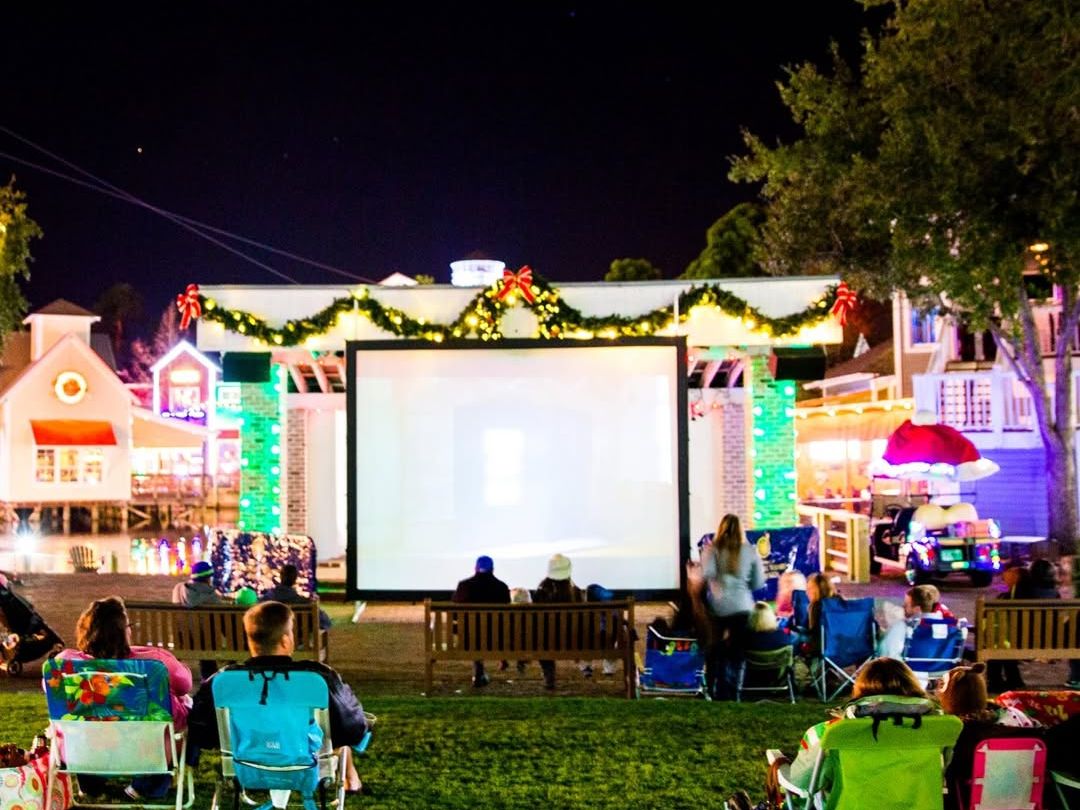 Families sit on lawn chairs and blankets while watching an outdoor holiday movie on a large screen at night. Festive lights, Christmas garlands, and nearby shops create a warm and cheerful setting for a community movie night. This image captures the cozy feel of outdoor holiday movies with family and friends.