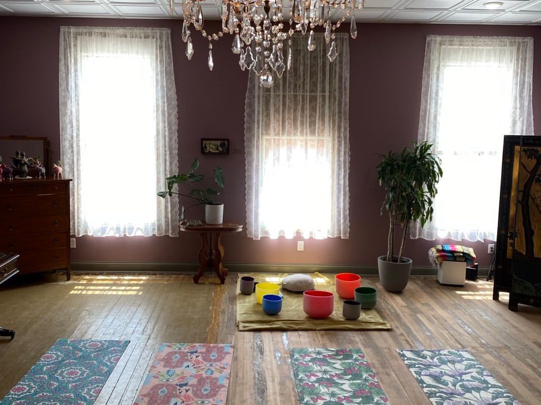 Yoga mats and crystal singing bowls are set up under a sparkling chandelier at Age of Aquarius Wellness. The bright room with lace curtains creates a peaceful space for sound healing and meditation.