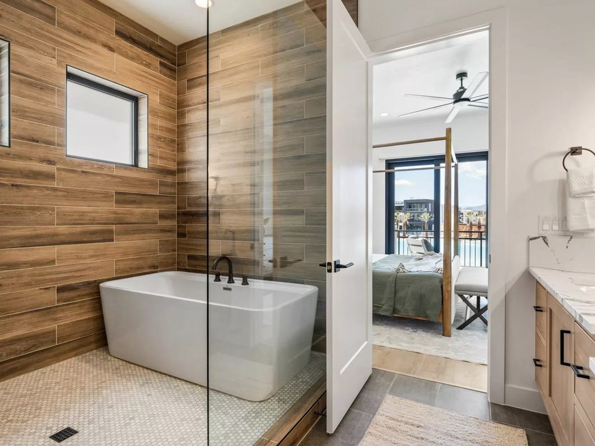 Modern bathroom with a wood-accented shower and soaking tub, leading into a bedroom with large windows and balcony views.