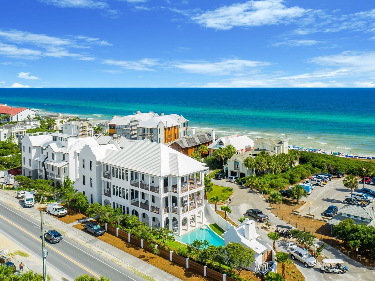 An aerial view of the Isle of Palms, South Carolina, shows a grand white multi-story beach house with a private pool steps from the turquoise Atlantic Ocean, surrounded by palm trees and neighboring coastal homes along the shoreline.