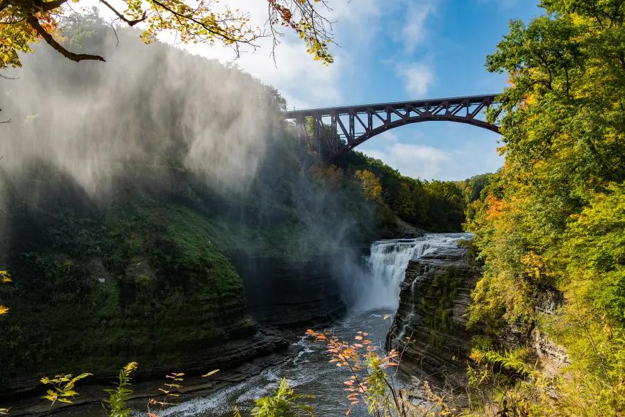 A wide view of Letchworth State Park showing a deep gorge, flowing waterfall, and the iconic bridge above, surrounded by fall colors. The mist rising from the falls adds drama to this scenic Finger Lakes landscape.