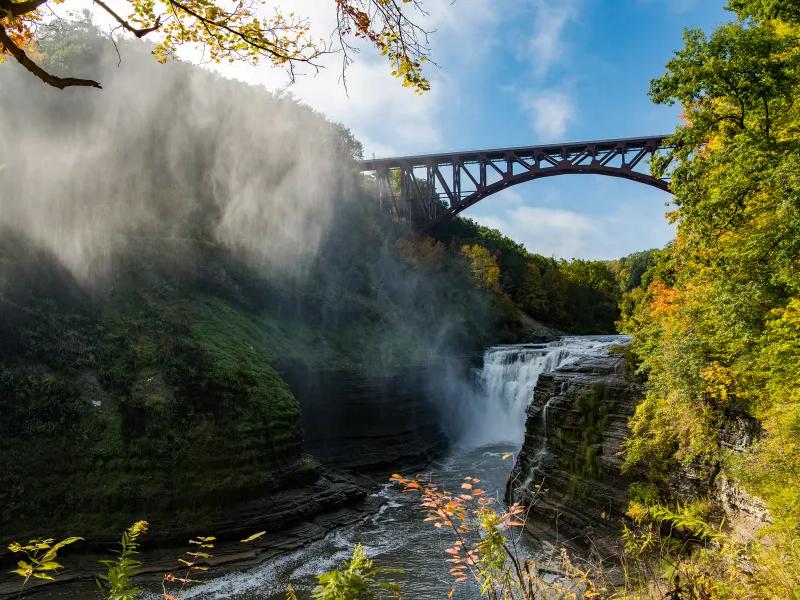 A wide view of Letchworth State Park showing a deep gorge, flowing waterfall, and the iconic bridge above, surrounded by fall colors. The mist rising from the falls adds drama to this scenic Finger Lakes landscape.