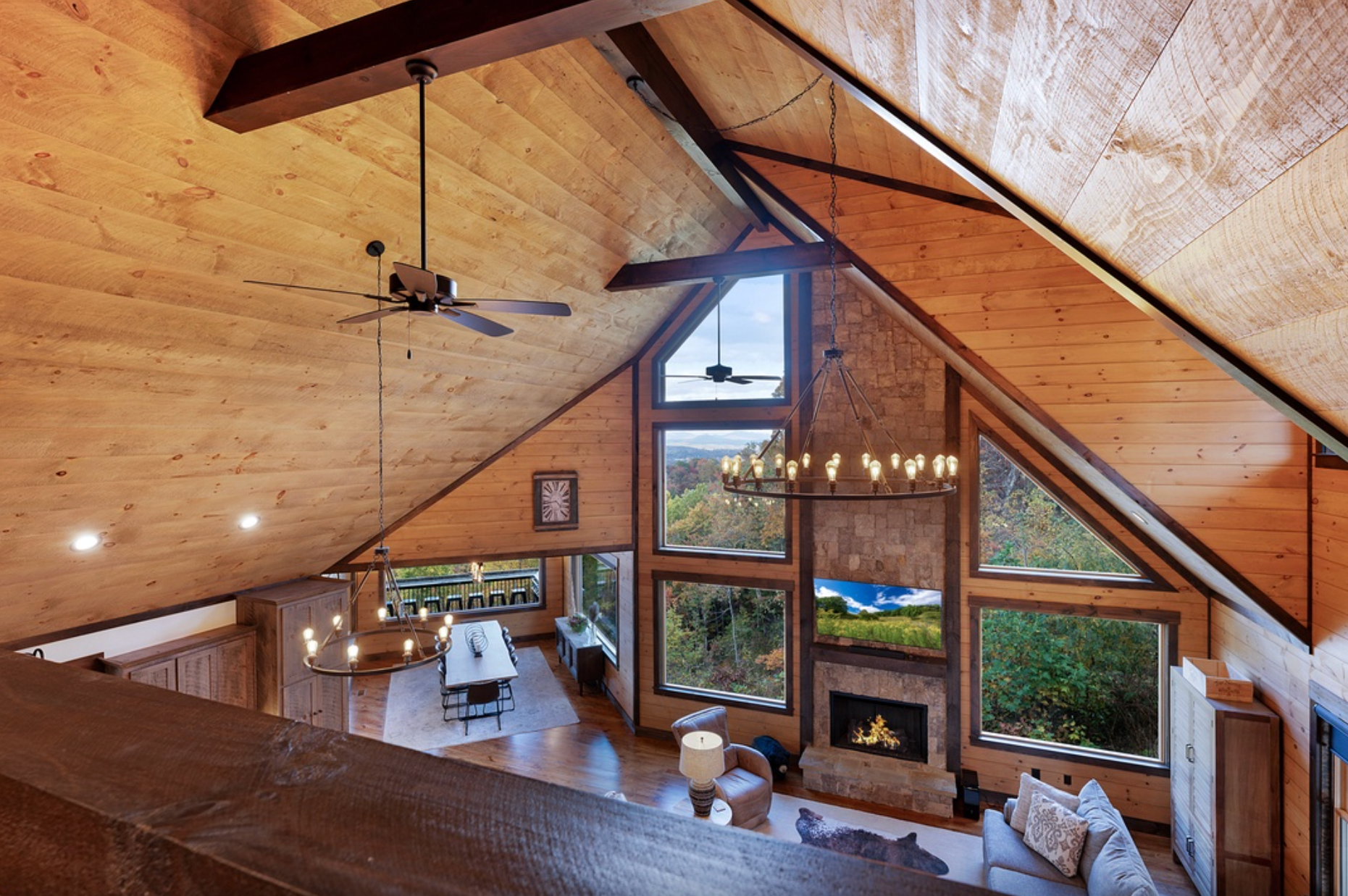 Vaulted wood-paneled great room with exposed beams, large triangular windows, chandelier lighting, and a stone fireplace overlooking a forested view.