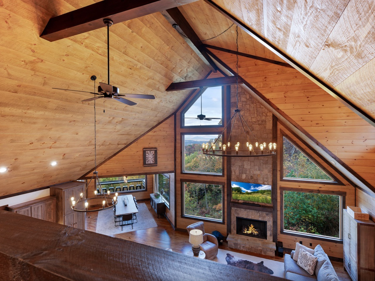 Vaulted wood-paneled great room with exposed beams, large triangular windows, chandelier lighting, and a stone fireplace overlooking a forested view.