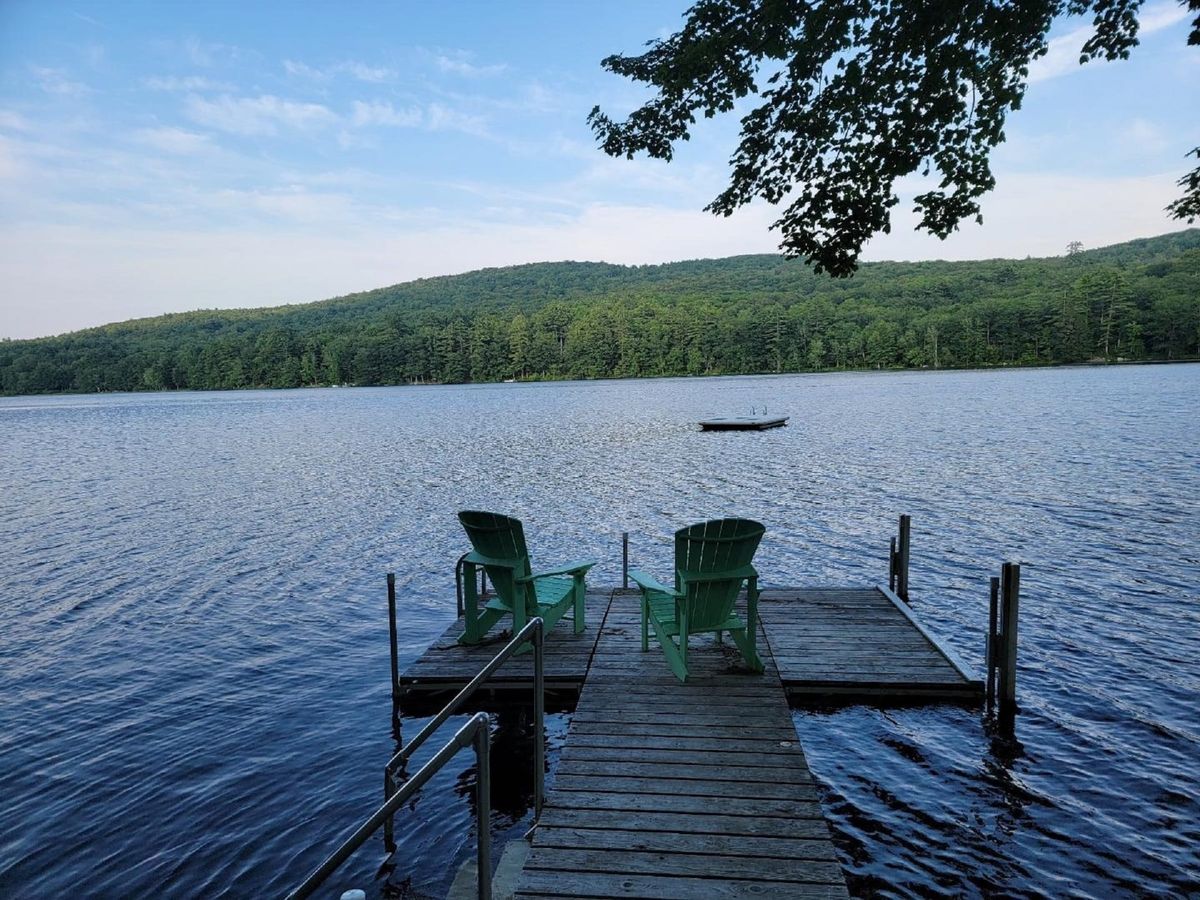 Two green Adirondack chairs sit at the end of a wooden dock overlooking a calm lake surrounded by forested hills under a blue sky.