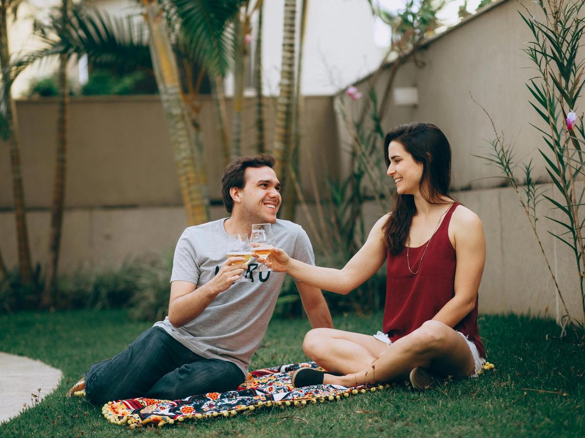 A smiling couple toasts with drinks while sitting on a blanket in a sunny backyard. The relaxed atmosphere reflects the personalized comfort and warmth of a private vacation rental stay.